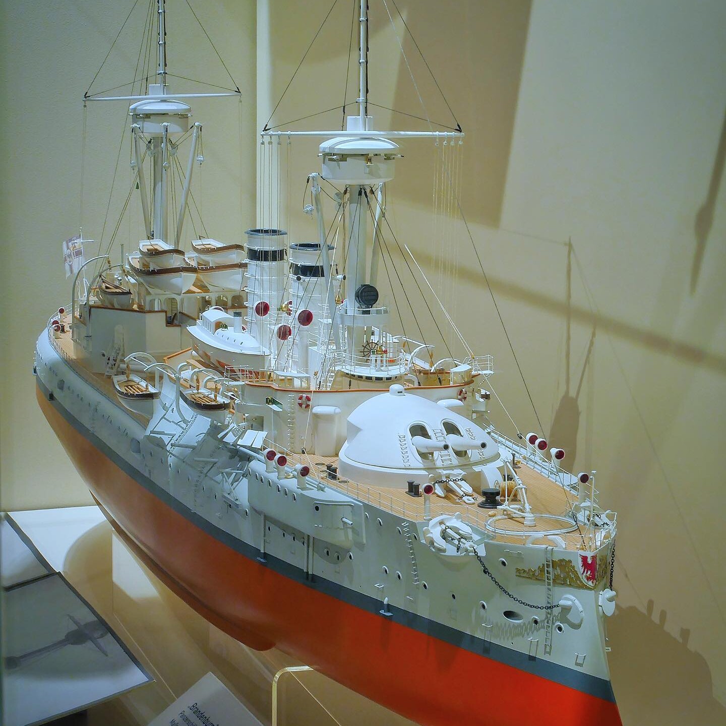 Detailliertes Modell eines Kriegsschiffs mit rotem Rumpf, weißen Aufbauten, Masten, Takelage und Rettungsbooten in einer Vitrine.