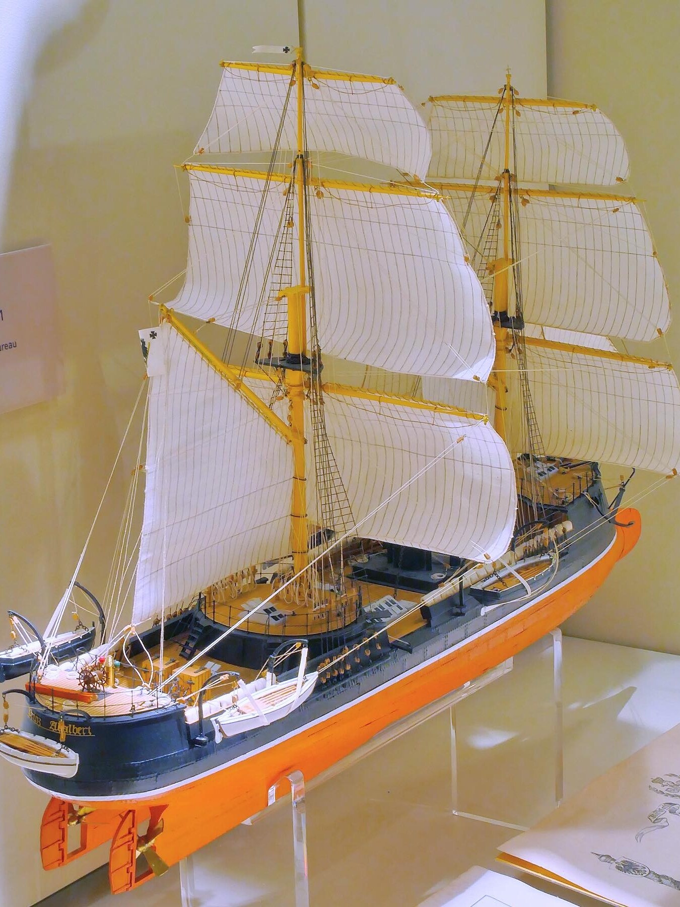 Detailliertes Schiffsmodell mit weißen geblähten Segeln, gelb gestrichenen Masten und orange-schwarzem Rumpf auf Ausstellungspodest.