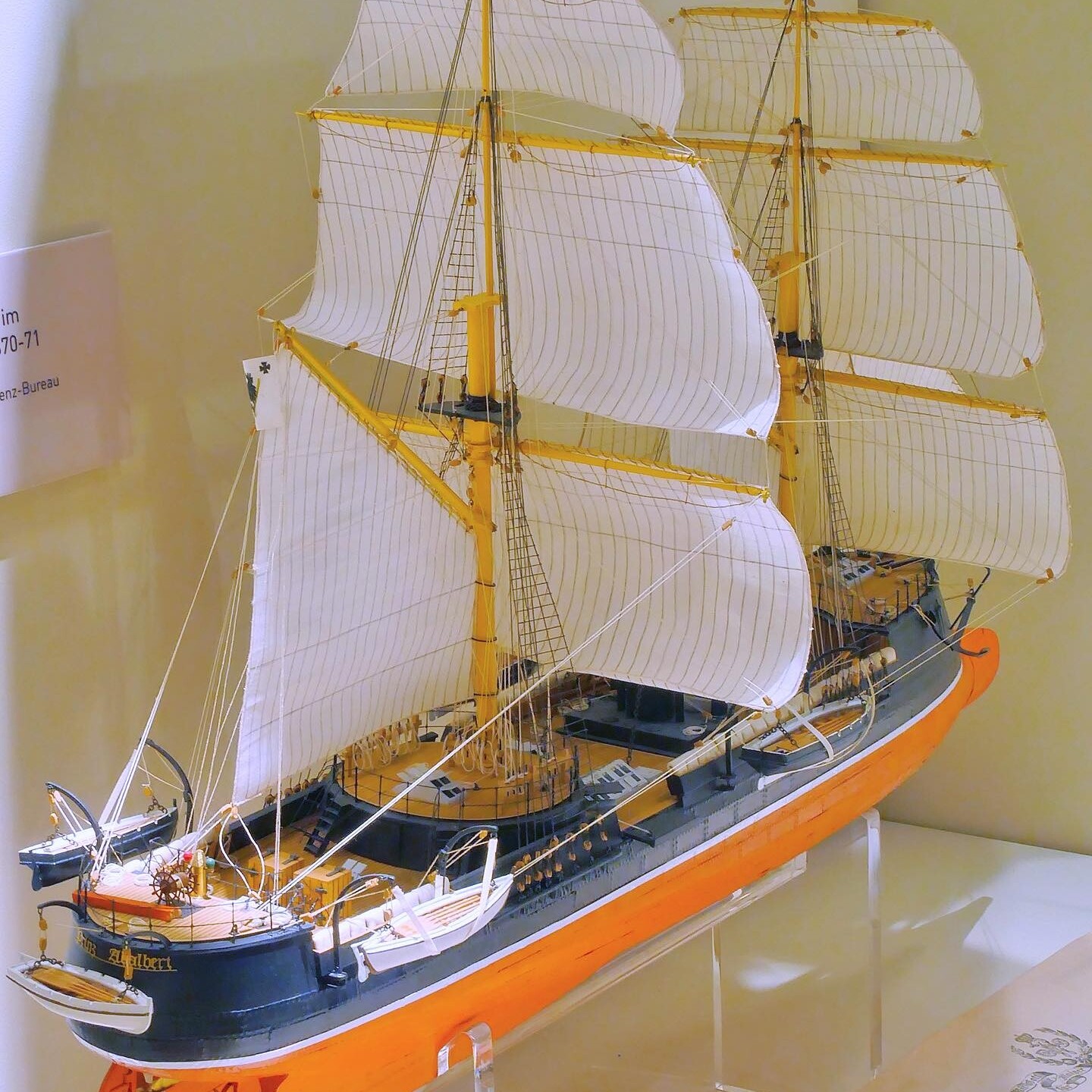Detailliertes Schiffsmodell mit weißen geblähten Segeln, gelb gestrichenen Masten und orange-schwarzem Rumpf auf Ausstellungspodest.