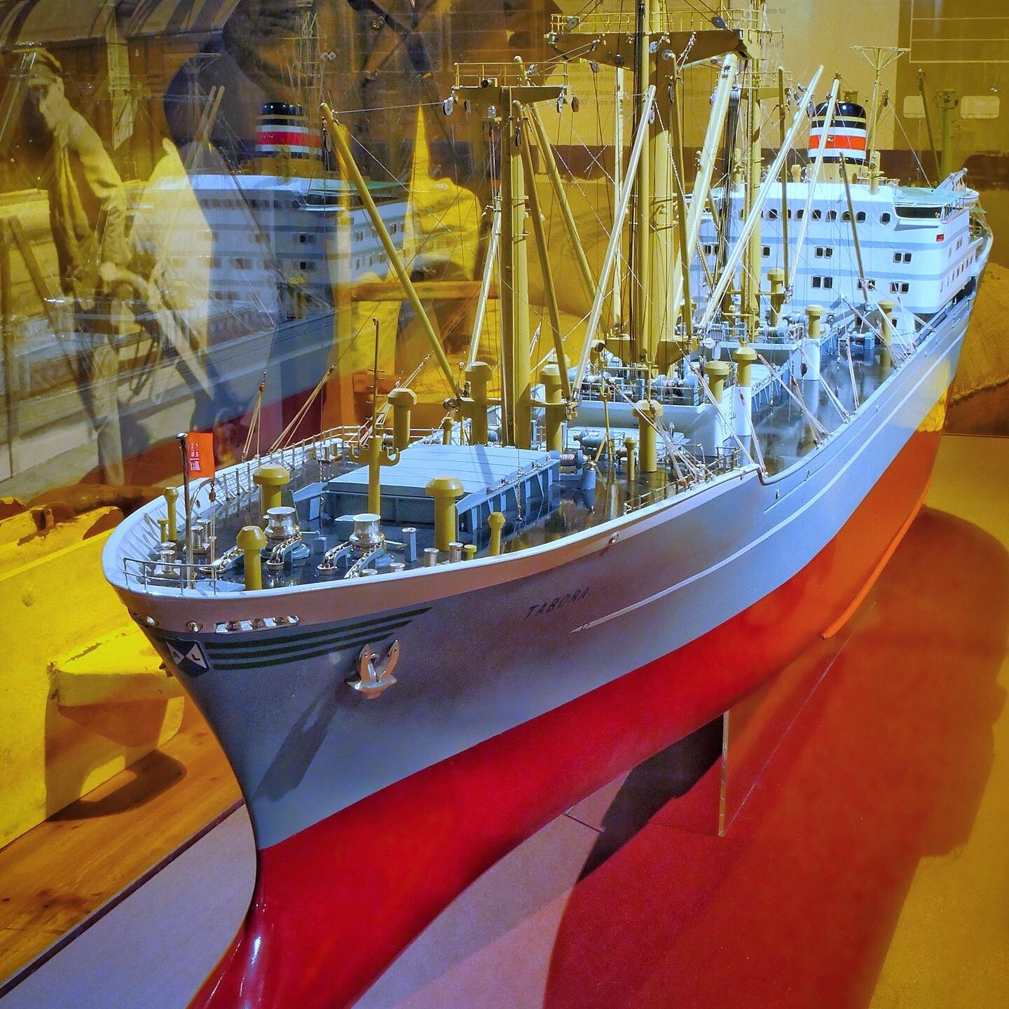 Detailliertes Modell eines Frachtschiffs mit rotem Rumpf, zahlreichen Ladebäumen, Takelage und Weißaufsatz, in Museumsvitrine mit Reflexionen.