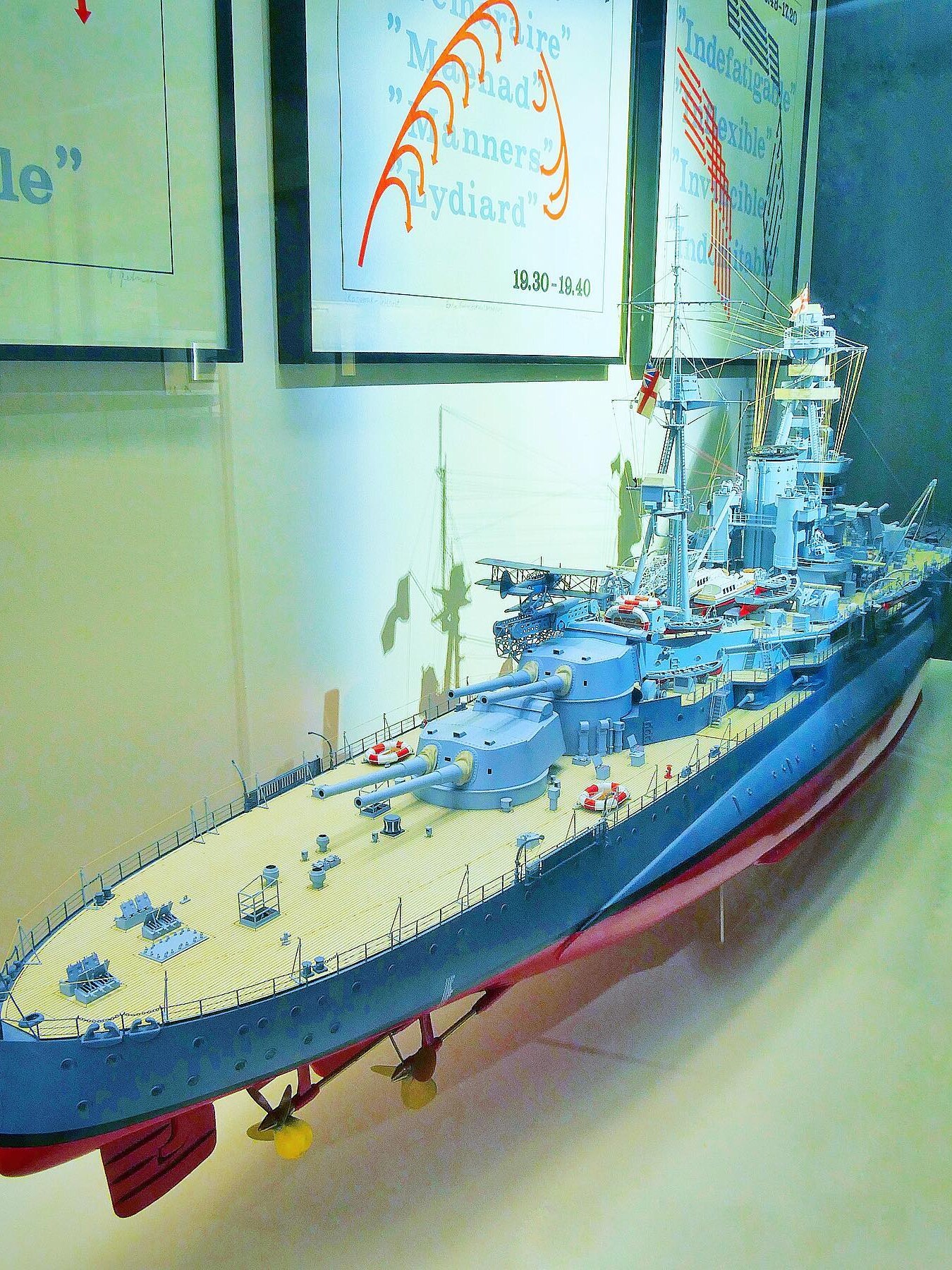 Detailliertes Modell eines graublauen Schlachtschiffs mit sichtbaren Kanonen, Deckseinrichtungen und Propellern, ausgestellt vor Plakaten.