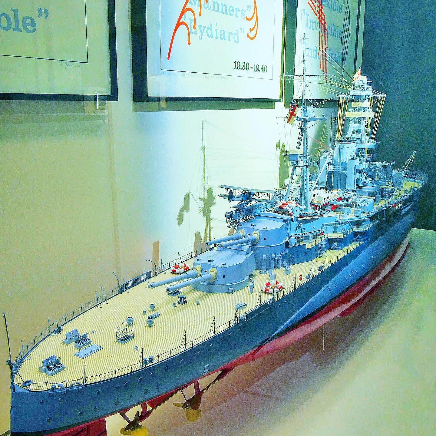 Detailliertes Modell eines graublauen Schlachtschiffs mit sichtbaren Kanonen, Deckseinrichtungen und Propellern, ausgestellt vor Plakaten.