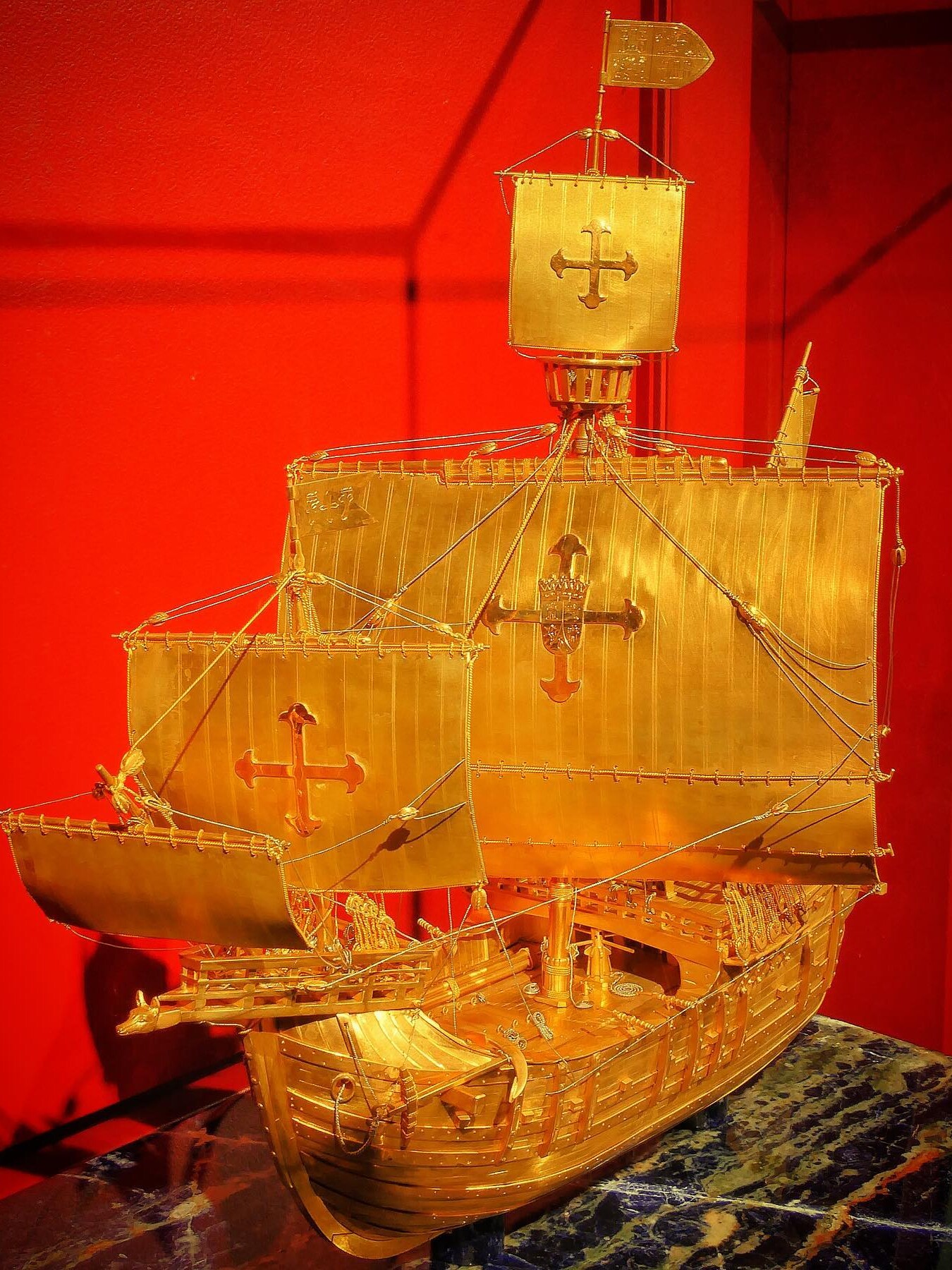 Goldenes Modell eines Segelschiffs mit kreuzförmigen Wappen auf den Segeln vor rotem Hintergrund.
