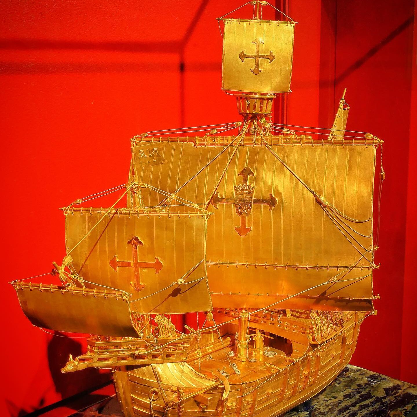 Goldenes Modell eines Segelschiffs mit kreuzförmigen Wappen auf den Segeln vor rotem Hintergrund.