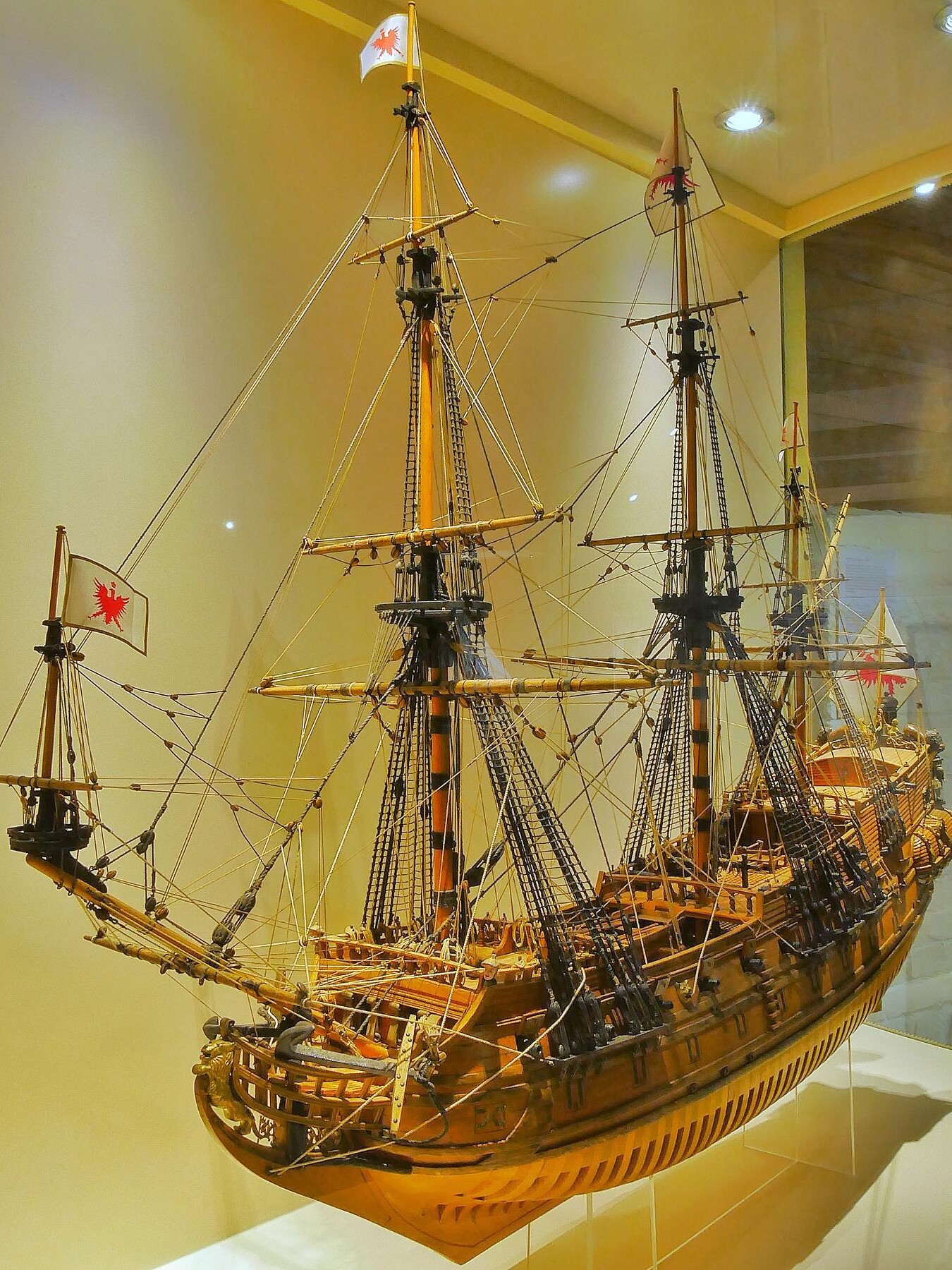 Detailliertes Holzmodell eines historischen Segelschiffs mit mehreren Masten, Takelagen und roten Wappenflaggen in einer Museumsvitrine.