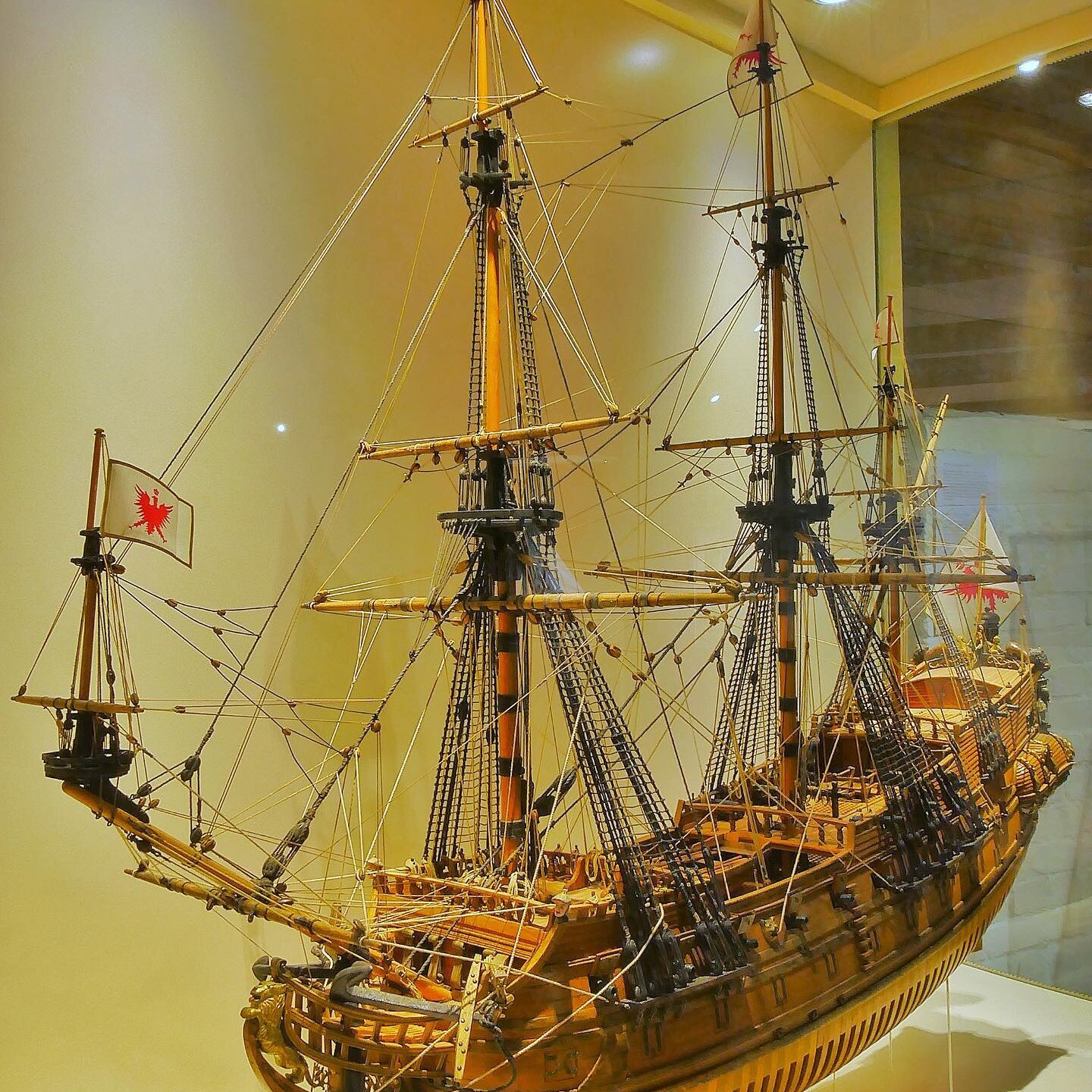 Detailliertes Holzmodell eines historischen Segelschiffs mit mehreren Masten, Takelagen und roten Wappenflaggen in einer Museumsvitrine.