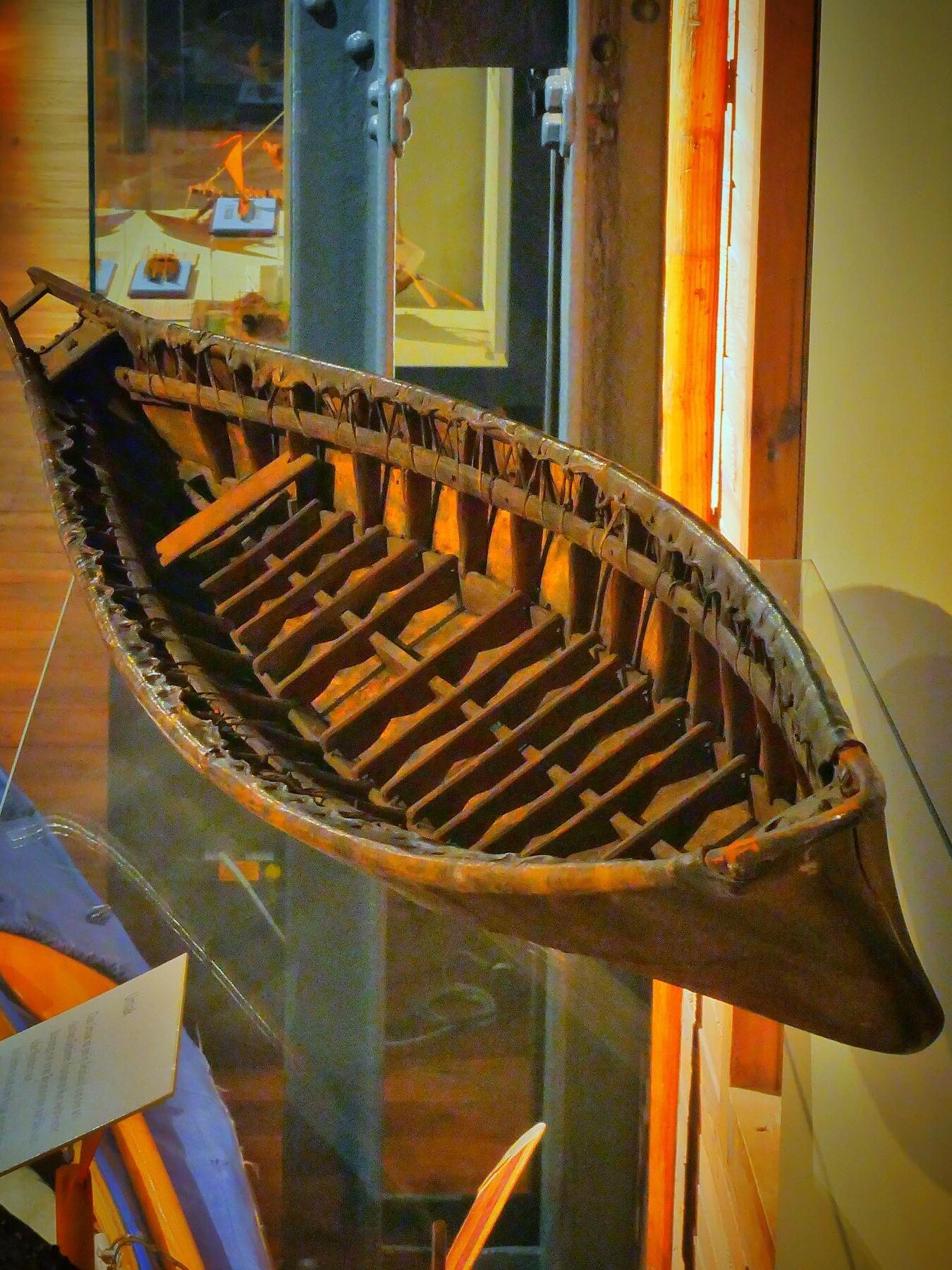 Halbe Ansicht eines alten, handgefertigten Holzkanus mit sichtbaren Rippen und Flechtverbindungen in Museumsvitrine.