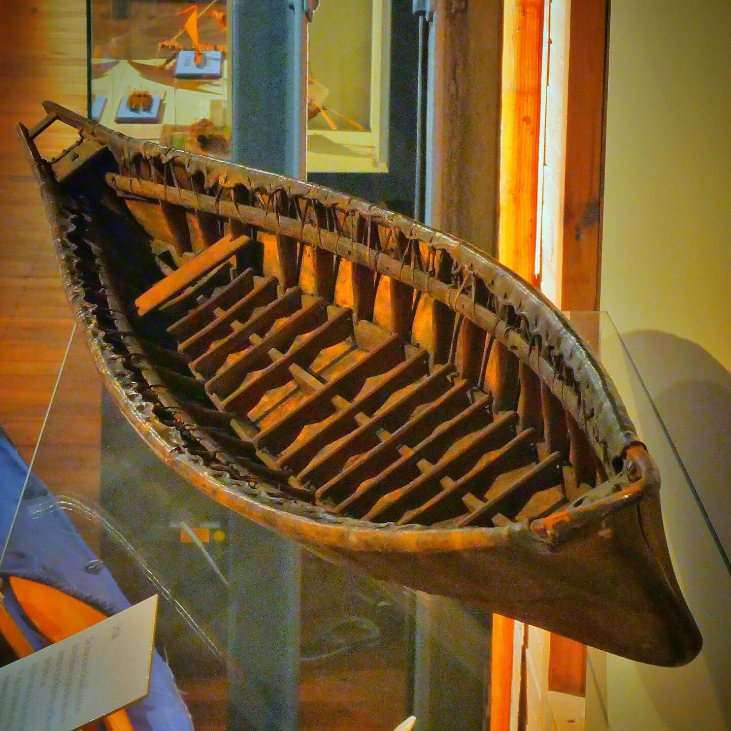 Halbe Ansicht eines alten, handgefertigten Holzkanus mit sichtbaren Rippen und Flechtverbindungen in Museumsvitrine.