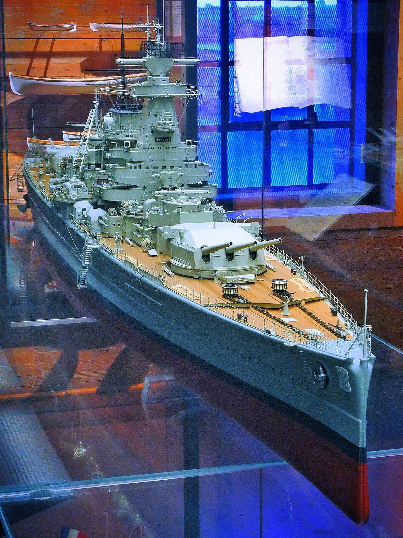 Detailliertes Schiffsmodell eines Schlachtschiffs mit vorderer Dreifachturm-Bewaffnung und sichtbarem Deck in einer Glasvitrine.