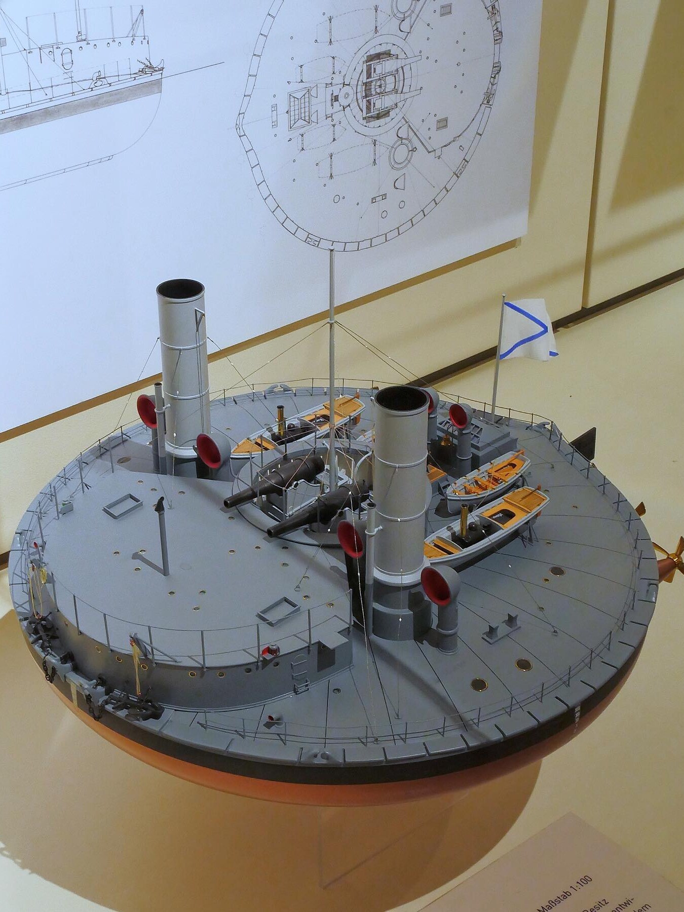 Rundes Schiffsmodell mit zwei Schornsteinen, Kanonen, Rettungsbooten und blau-weißem Flaggenmast vor technischen Zeichnungen.