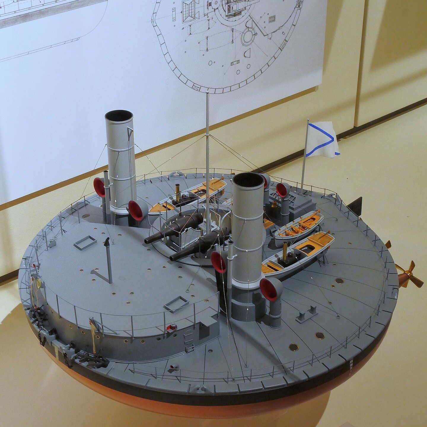 Rundes Schiffsmodell mit zwei Schornsteinen, Kanonen, Rettungsbooten und blau-weißem Flaggenmast vor technischen Zeichnungen.