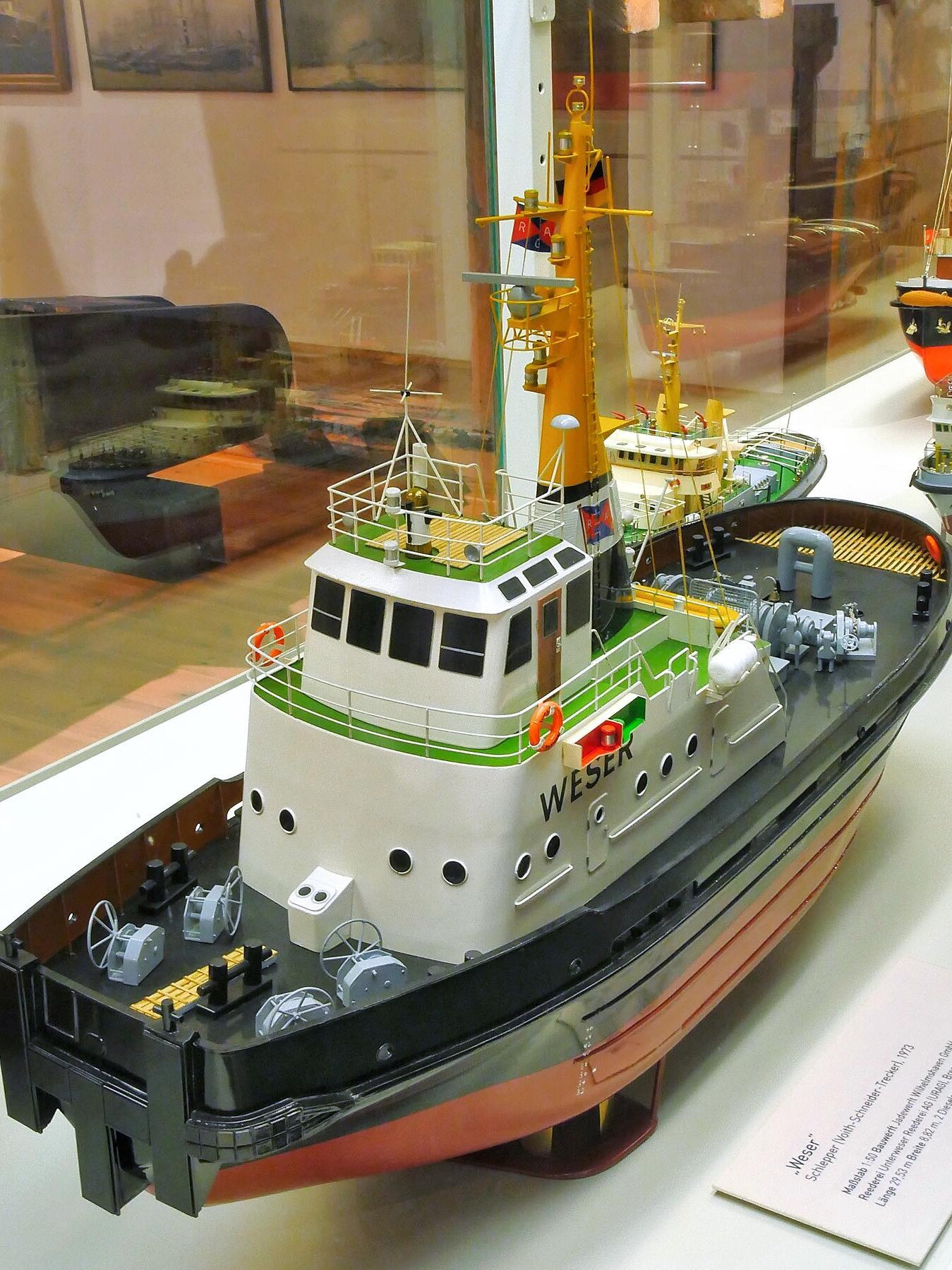 Detailliertes Modell eines Schlepperboots namens „Weser“ mit gelbem Mast, roten Rettungsringen und schwarzen Rumpf in einer Vitrine.