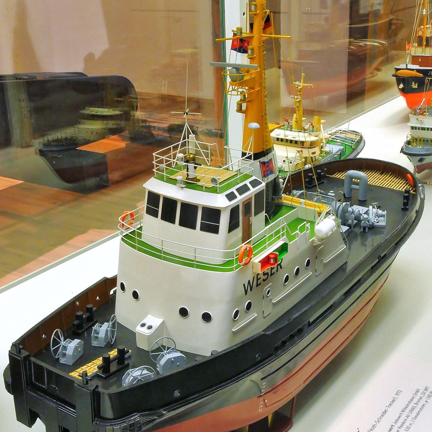 Detailliertes Modell eines Schlepperboots namens „Weser“ mit gelbem Mast, roten Rettungsringen und schwarzen Rumpf in einer Vitrine.