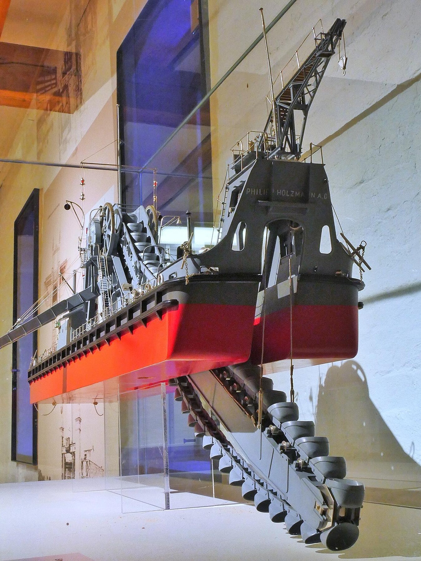 Modell eines Baggerbohrschiffs mit rot-schwarzem Rumpf, langem Kettenförderer und hoher Deckskran in Museumsausstellung.