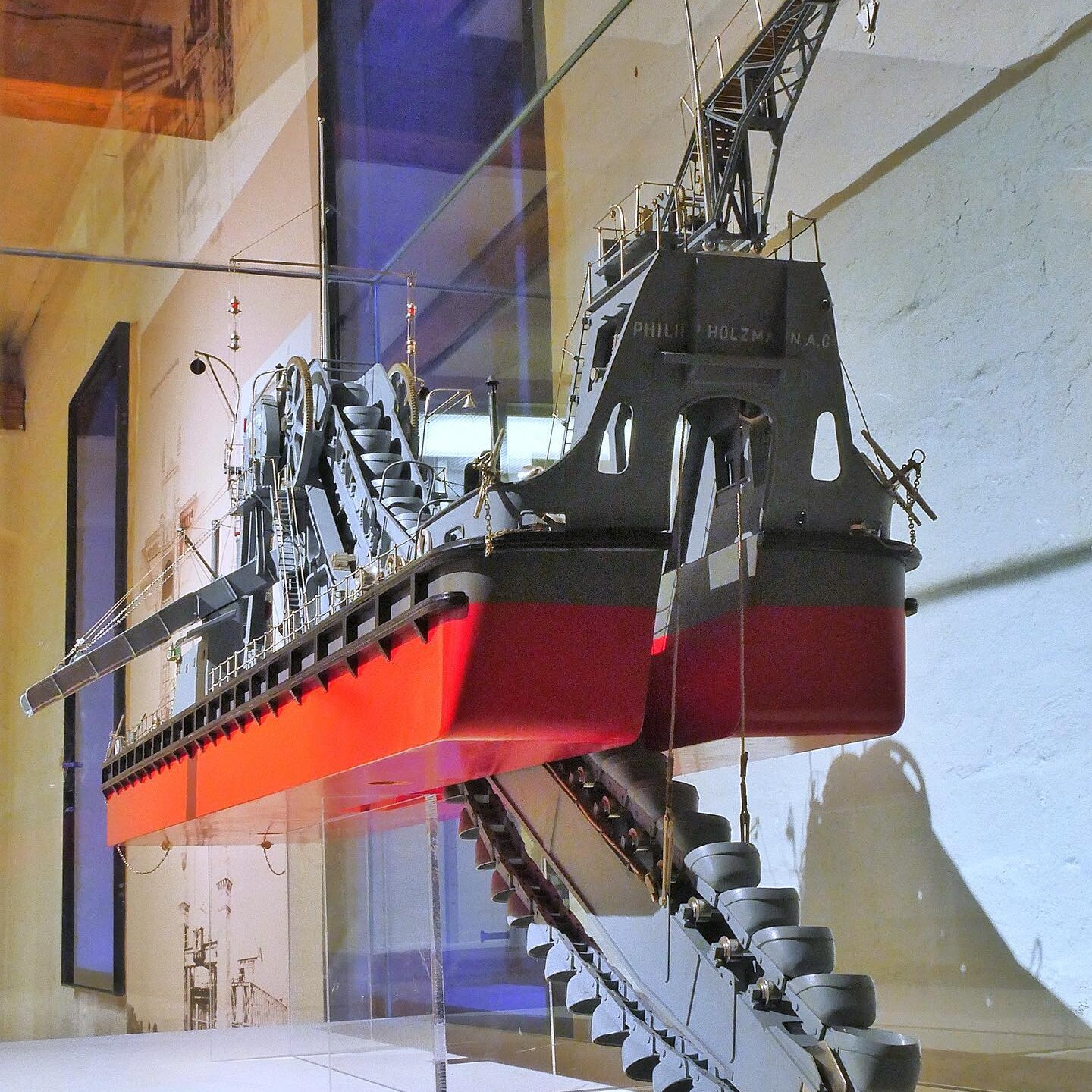 Modell eines Baggerbohrschiffs mit rot-schwarzem Rumpf, langem Kettenförderer und hoher Deckskran in Museumsausstellung.