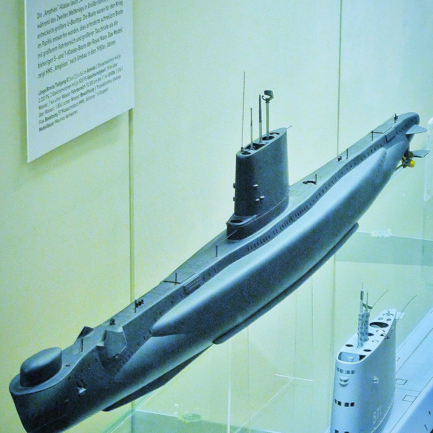 Modell eines grauen U-Boot-Rumpfes mit Turm, Antennen und Schraube in einer Vitrine, daneben Beschreibungszettel an der Wand