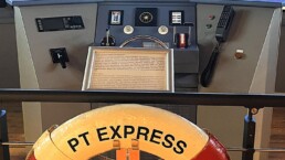 Rettungsring mit Aufschrift PT EXPRESS HAMBURG vor Steuerrad und mehreren Monitoren einer Schiffsbrücke.