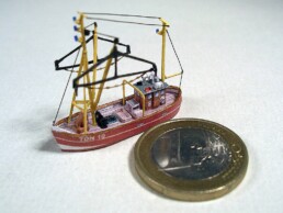 winziges rotes Modellschiff mit gelben Masten und schwarzem Ausleger neben einer Ein-Euro-Münze zur Größendarstellung