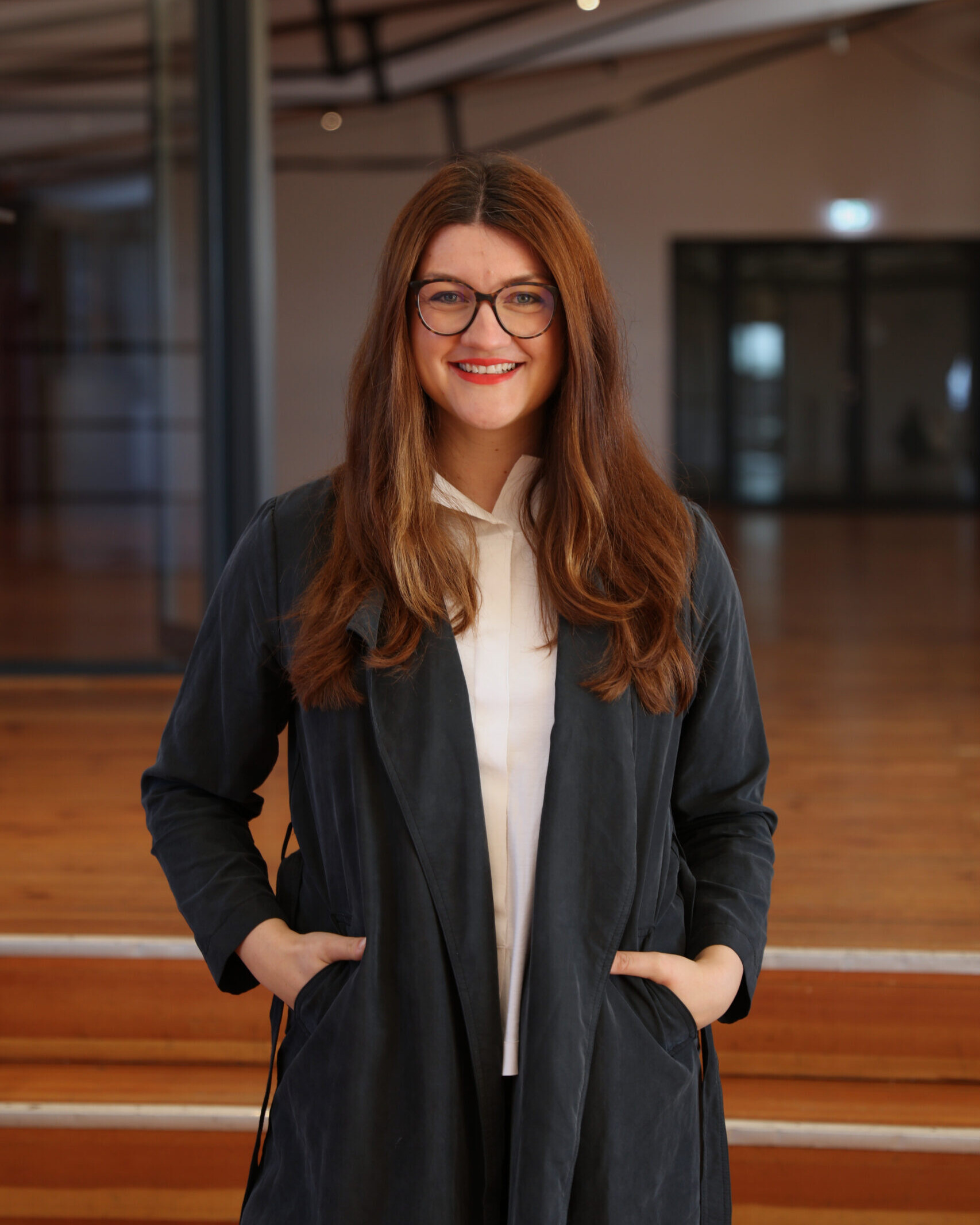 Frau mit langen braunen Haaren und Brille, lächelnd in dunklem Mantel und weißer Bluse vor Holzstufen.