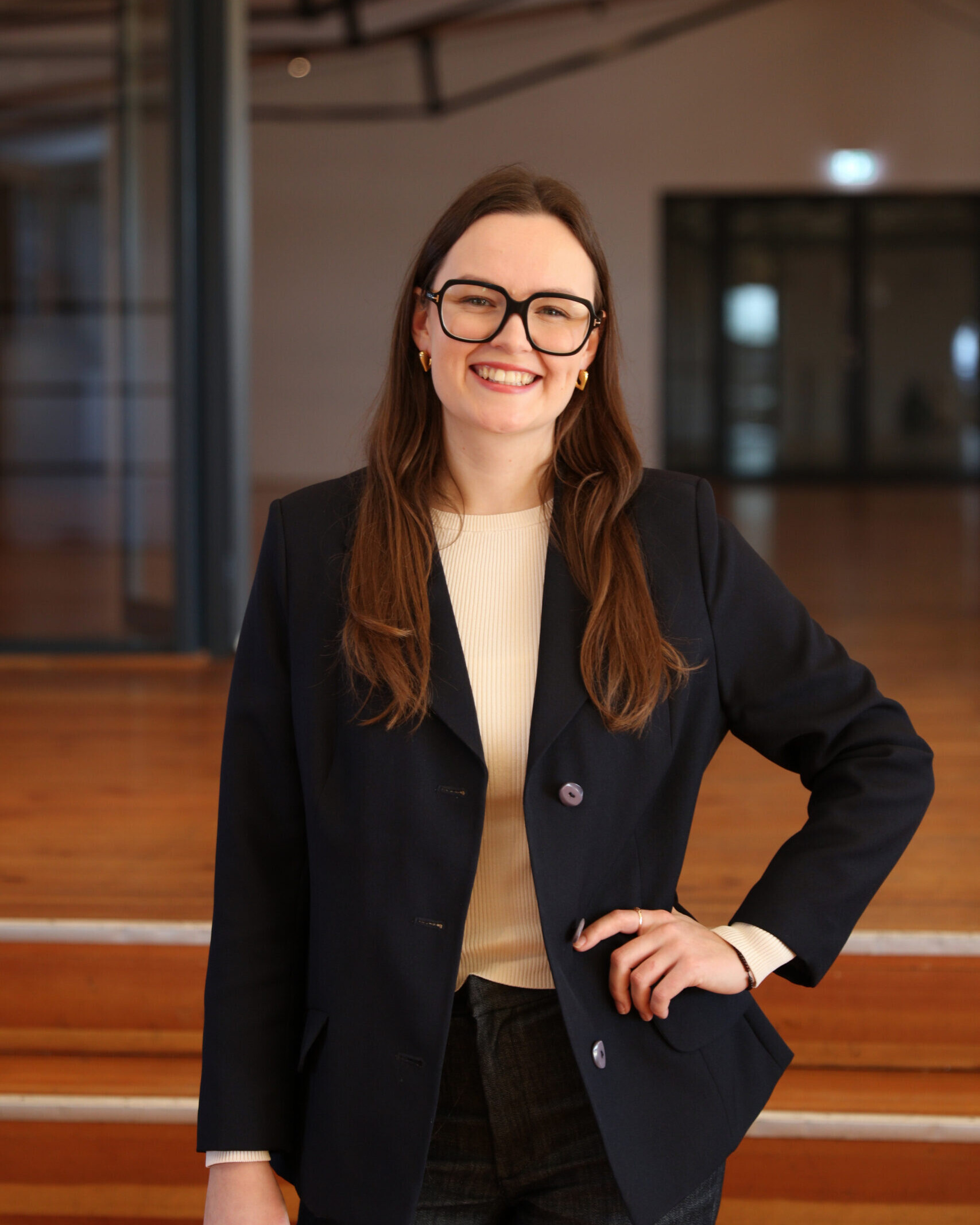 Lächelnde Frau mit schwarzer Brille, langen braunen Haaren und dunkelblauem Blazer, mit einer Hand in der Hüfte vor Holzstufen.