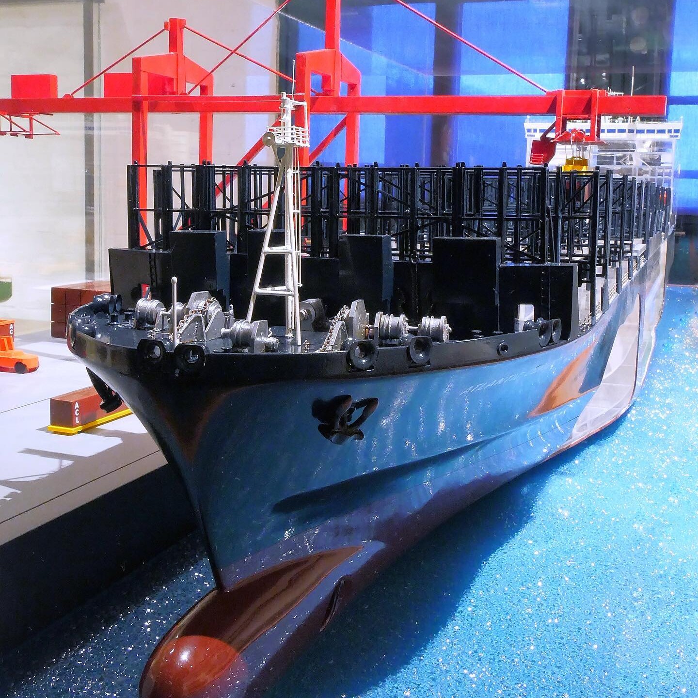 Modell eines Frachtschiffs mit blauem Rumpf, schwarzem Containergestell und rotem Hafenkran im Hintergrund.
