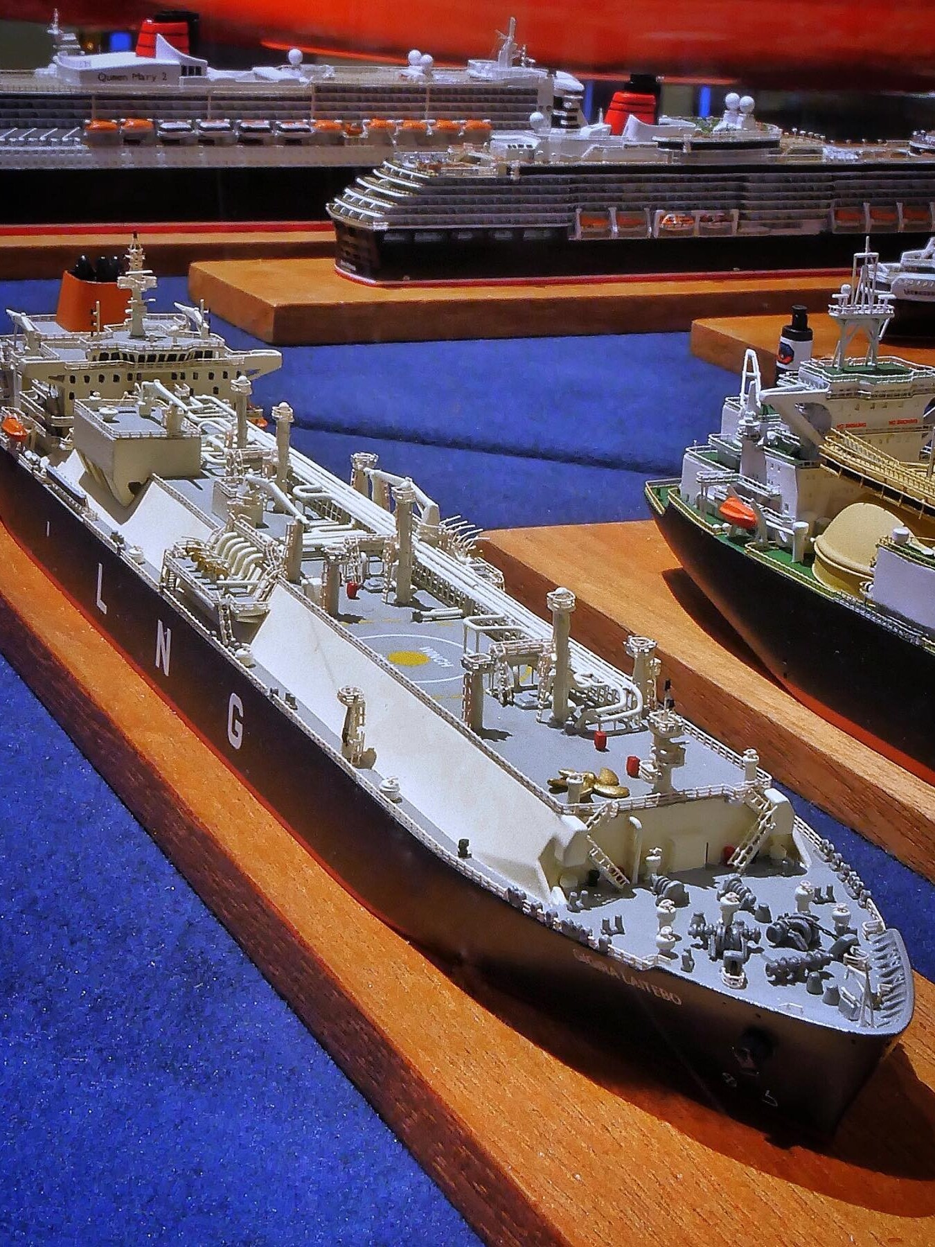 Mehrere detaillierte Schiffsmodelle auf Holzsockeln, vorn ein grauer Tankschiff-Prototyp mit Decksleitungen und Kräne, im Hintergrund Kreuzfahrtschiffe.