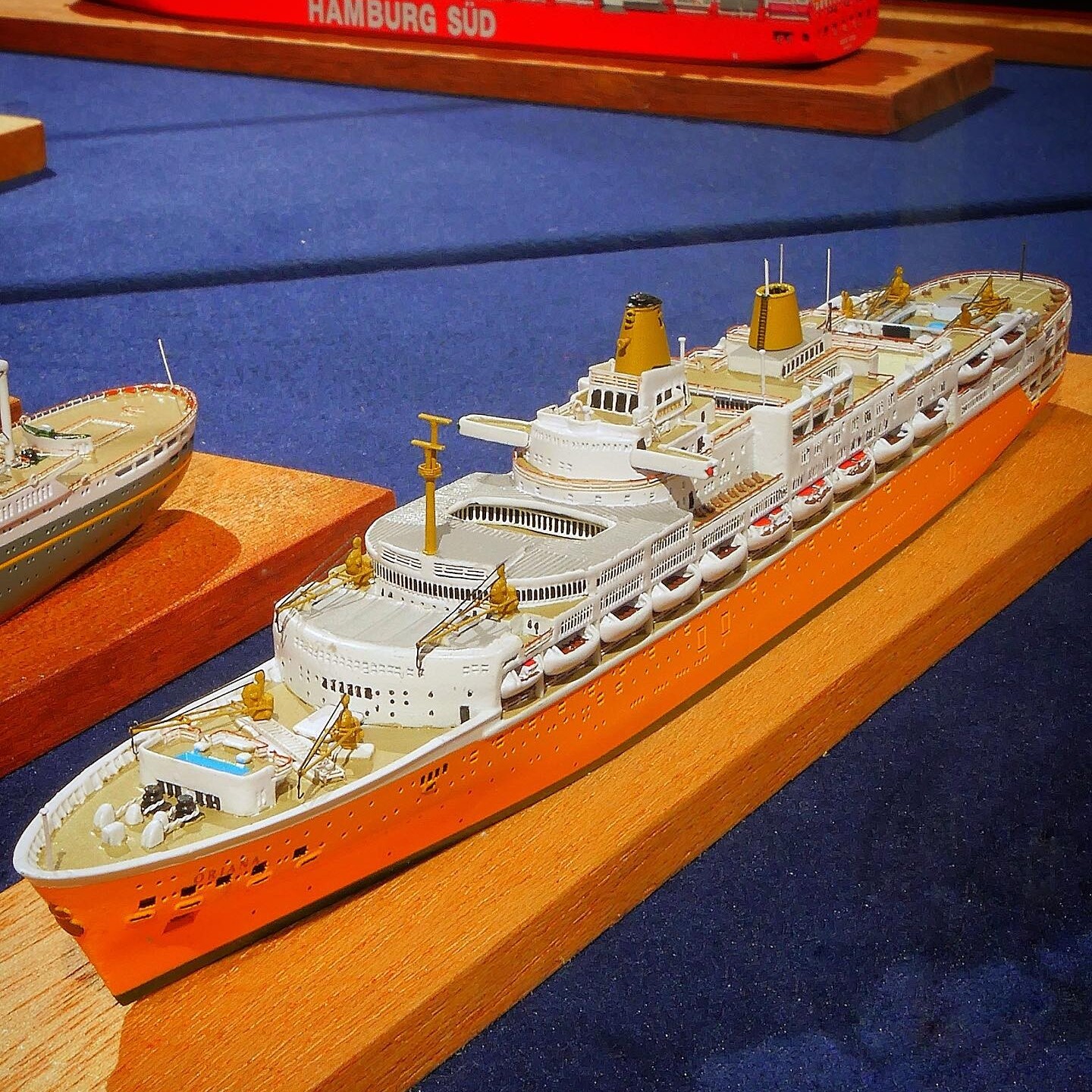 Modell eines orange-weißen Passagierschiffs mit Rettungsbooten und Deckdetails auf Holzsockel, im Hintergrund rotes Containerschiff mit Aufschrift HAMBURG SÜD.