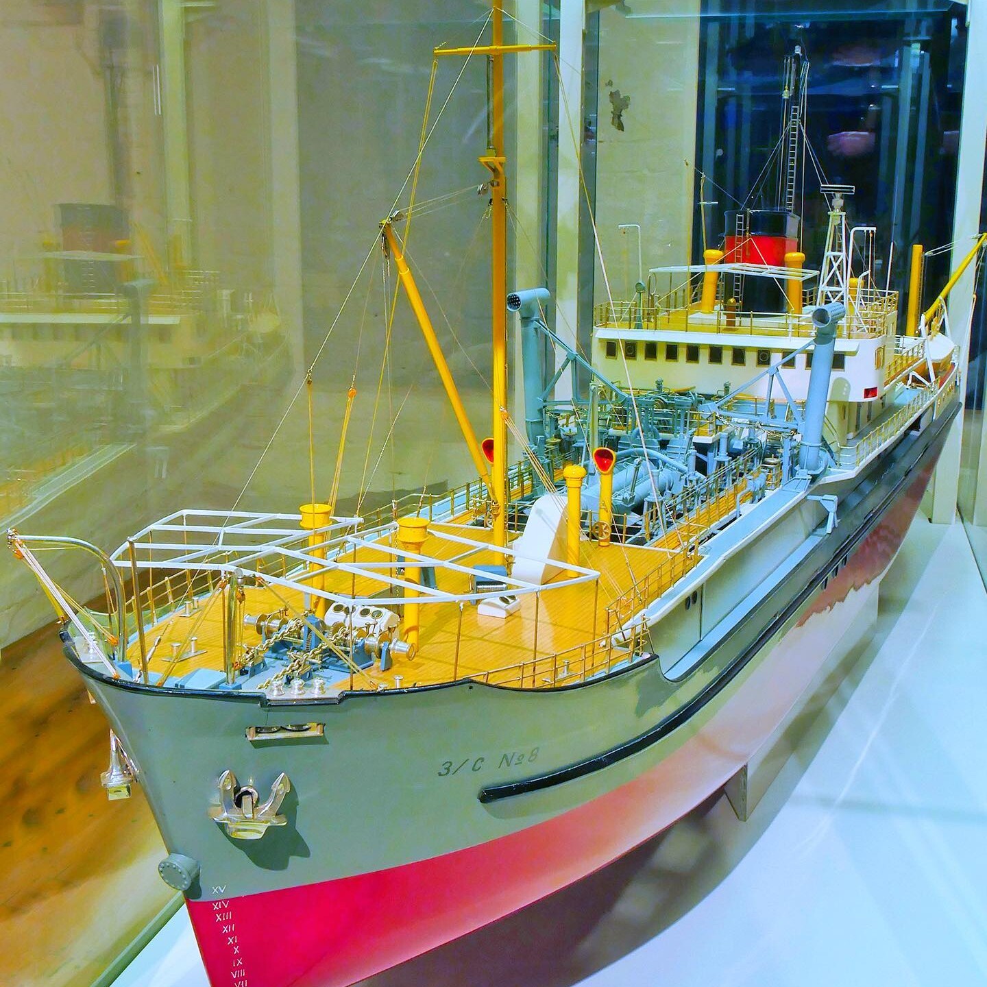 Detailliertes Schiffsmodell in Vitrine mit gelbem Deck, Masten, Takelage und rotem Unterwasserbereich, von schräg vorn gezeigt.