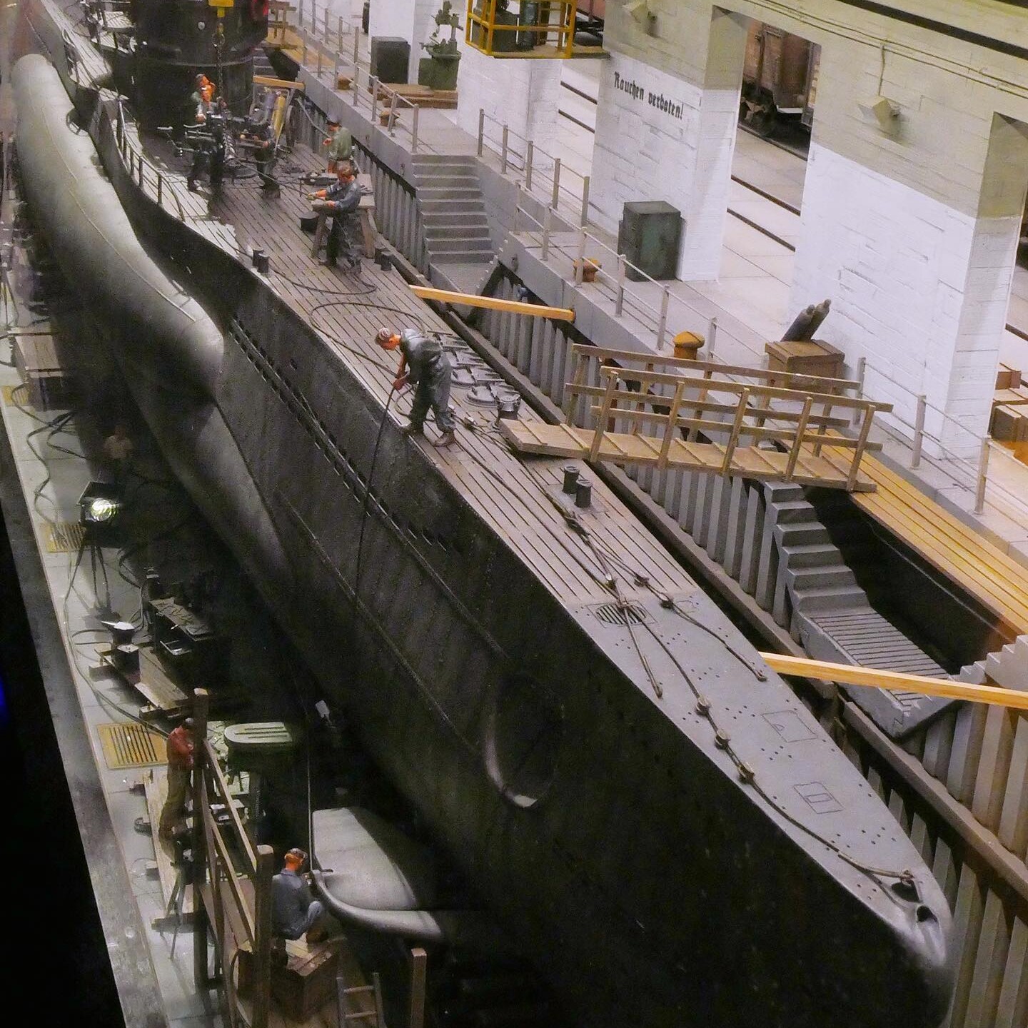 U-Boot im Trockendock mit Arbeitern auf dem Deck und Gerüst, industrielle Halle mit Geländern und Kranbahn im Hintergrund