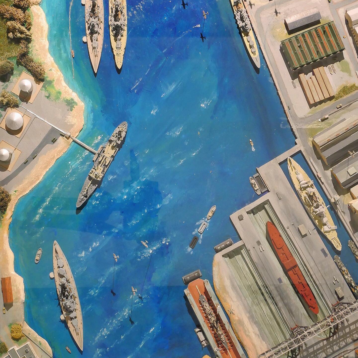 Luftaufnahme eines Hafens mit Kriegsschiffen, Frachtschiffen, Docks und kleinen Booten auf tiefblauem Wasser, umliegende Lagerhallen und Tankbehälter.