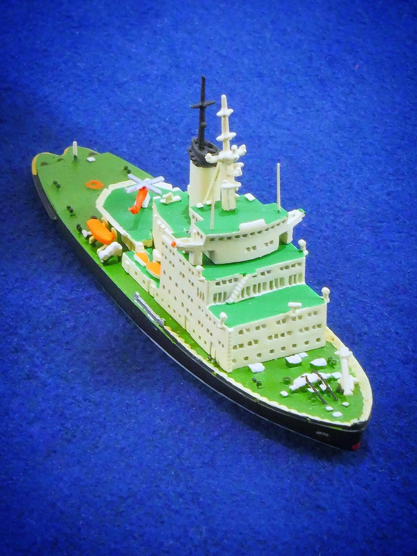 kleines Modellschiff mit grünen Decks, weißen Aufbauten und orangefarbenen Rettungsbooten auf blauem Untergrund, schräg von vorn aufgenommen