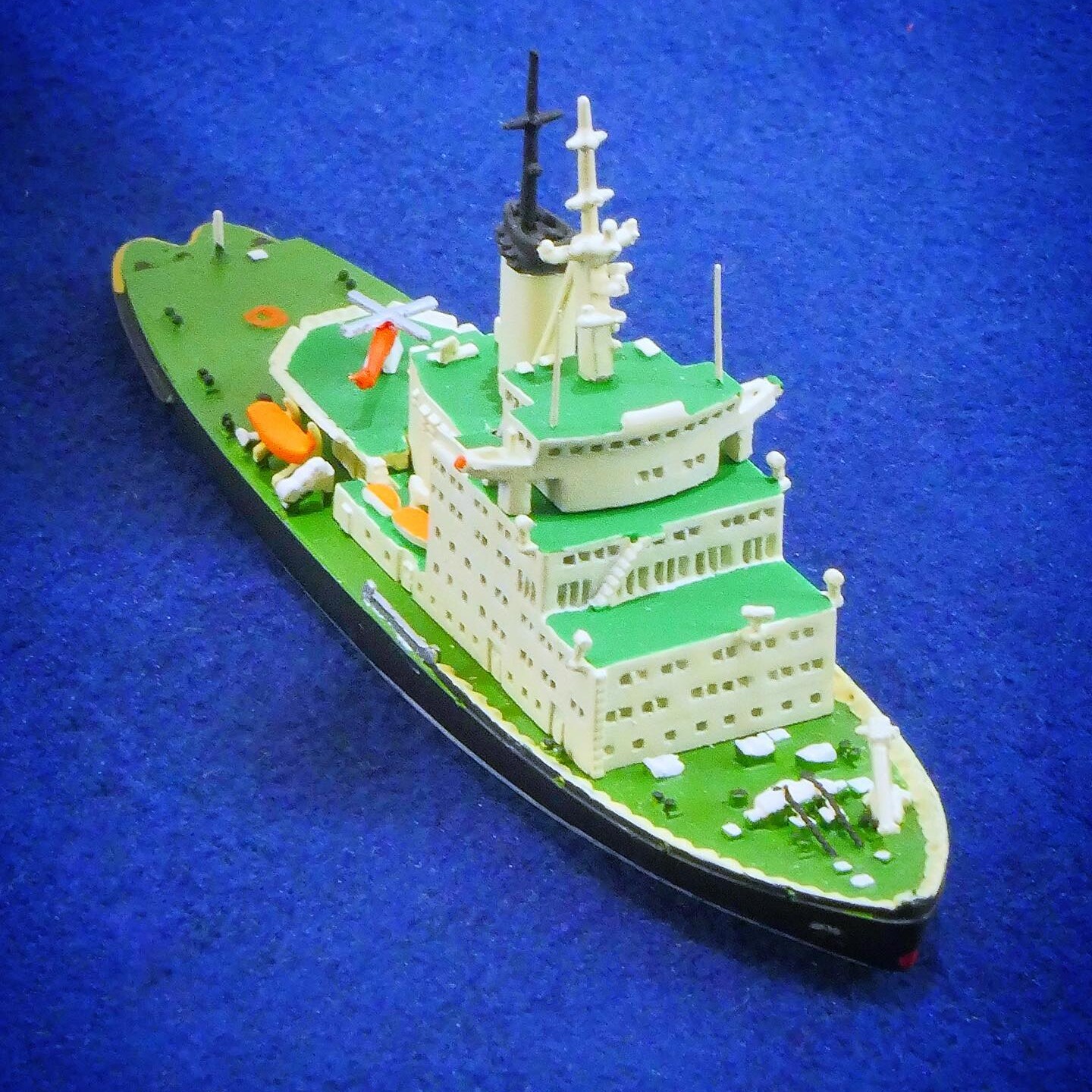 kleines Modellschiff mit grünen Decks, weißen Aufbauten und orangefarbenen Rettungsbooten auf blauem Untergrund, schräg von vorn aufgenommen