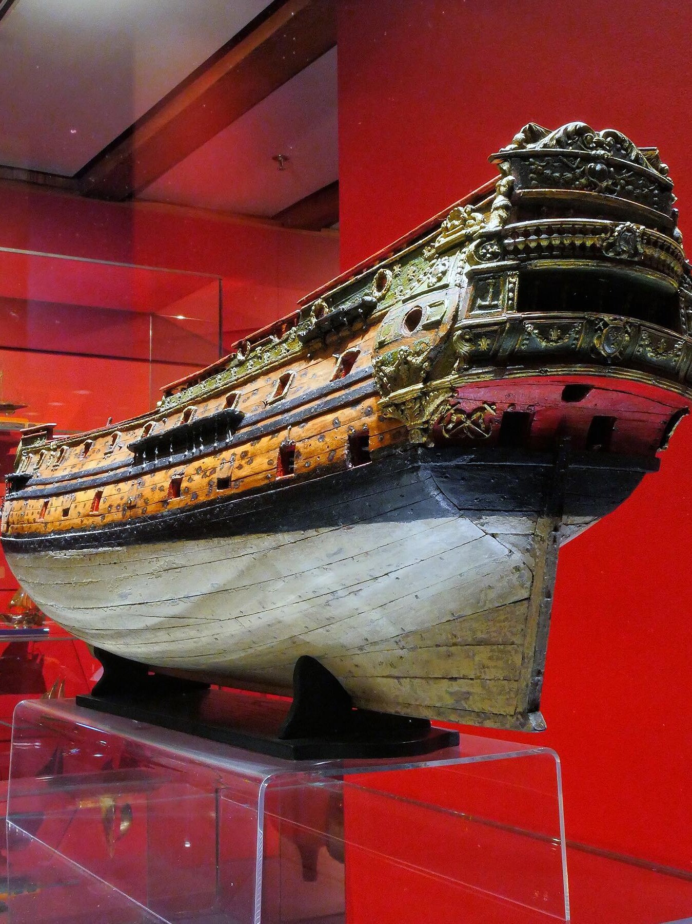 Holzmodell eines prunkvollen Segelschiffs mit vergoldeten Schnitzereien am Heck, auf rotem Museumsdisplay.