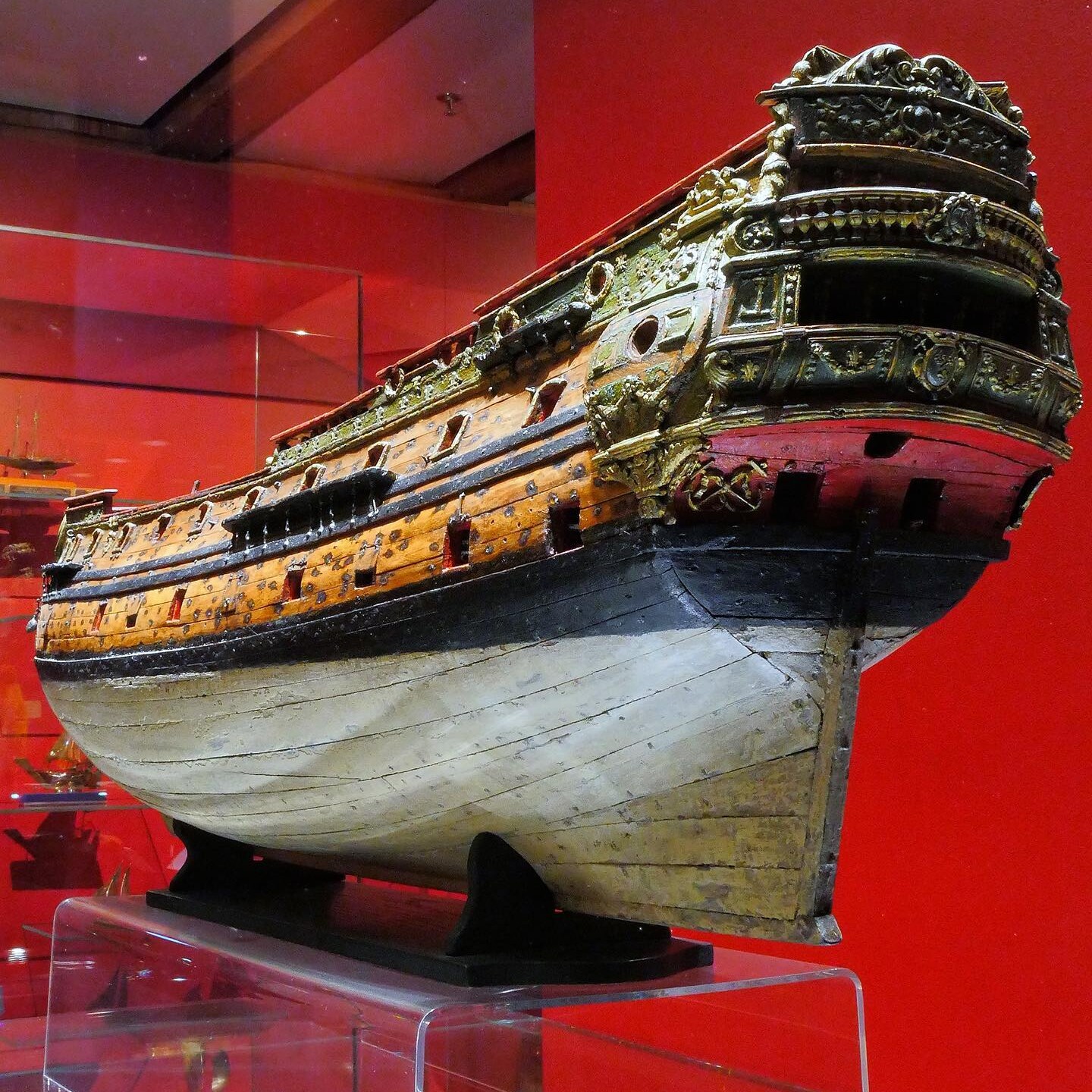 Holzmodell eines prunkvollen Segelschiffs mit vergoldeten Schnitzereien am Heck, auf rotem Museumsdisplay.