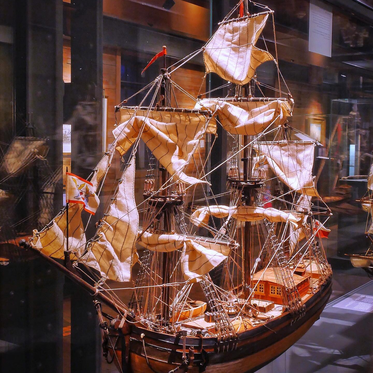 Detailliertes hölzernes Schiffsmodell mit geblähten Segeln, Takelage und kleinen Flaggen in einer Museumsvitrine.