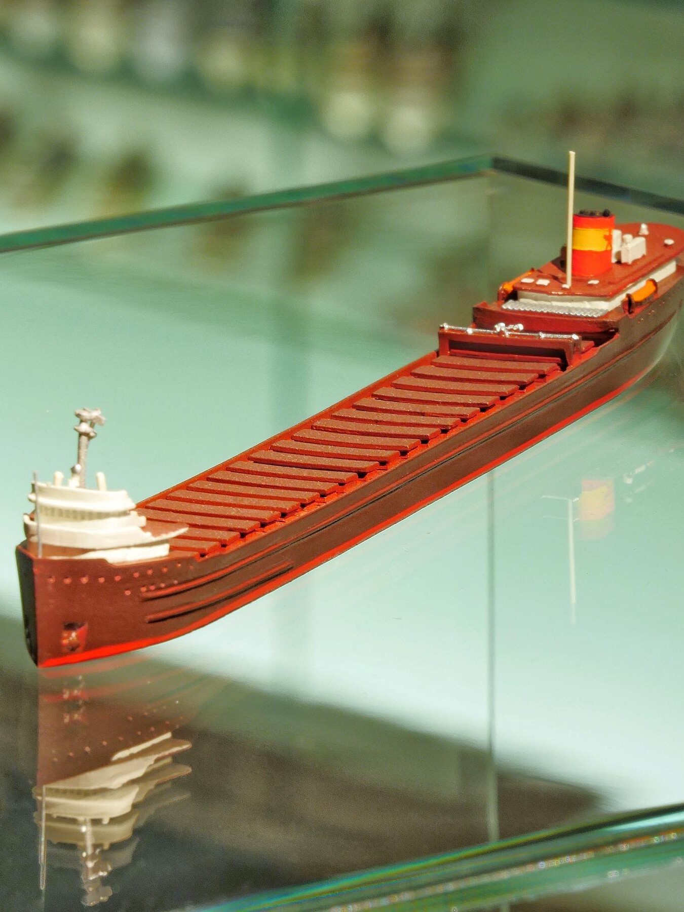 Rotes Modellfrachtschiff mit aufgesetzter Brücke vorne und Schornstein hinten, auf glänzender Glasfläche mit Spiegelung.