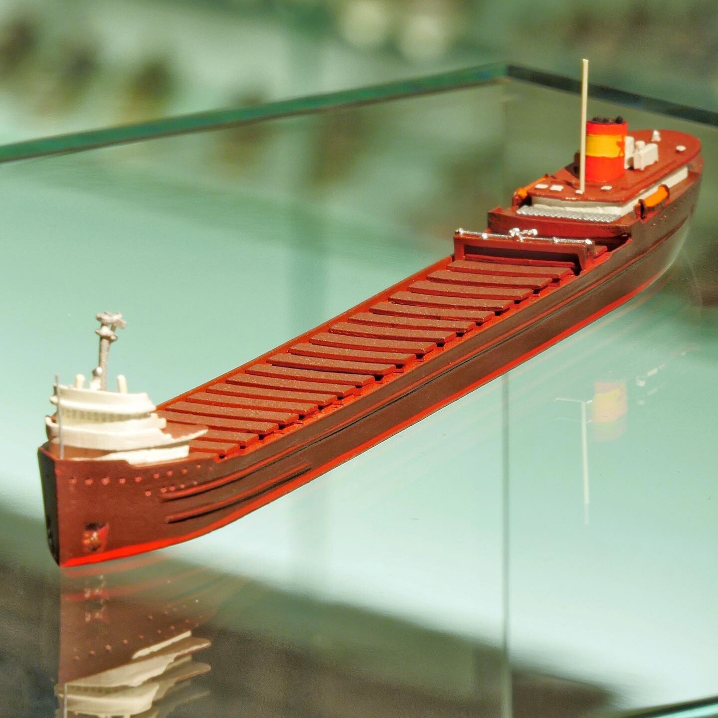 Rotes Modellfrachtschiff mit aufgesetzter Brücke vorne und Schornstein hinten, auf glänzender Glasfläche mit Spiegelung.