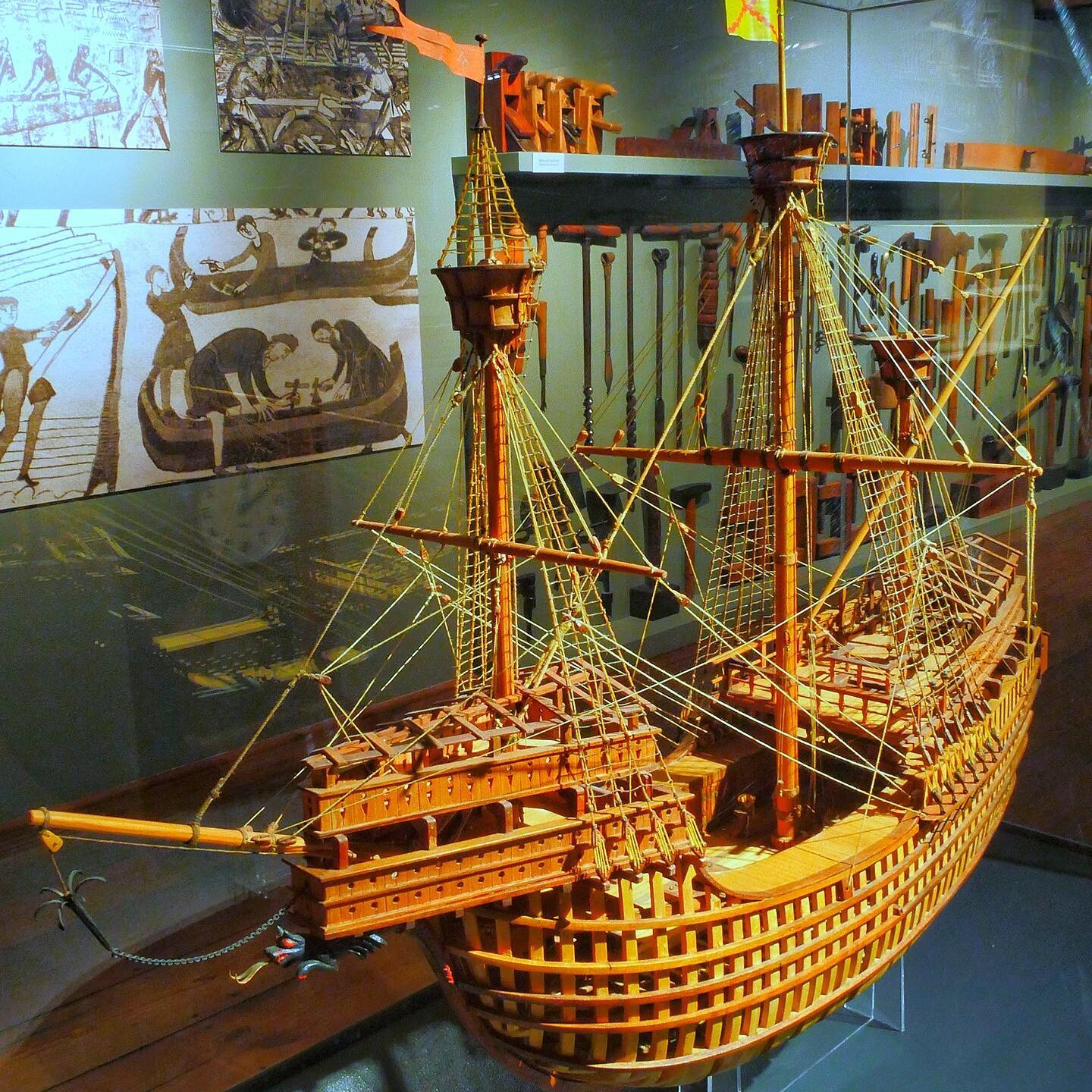 Holzmodell eines großen Segelschiffs mit Masten, Takelage und aufgesetzten Kastellen, ausgestellt vor historischen Zeichnungen.