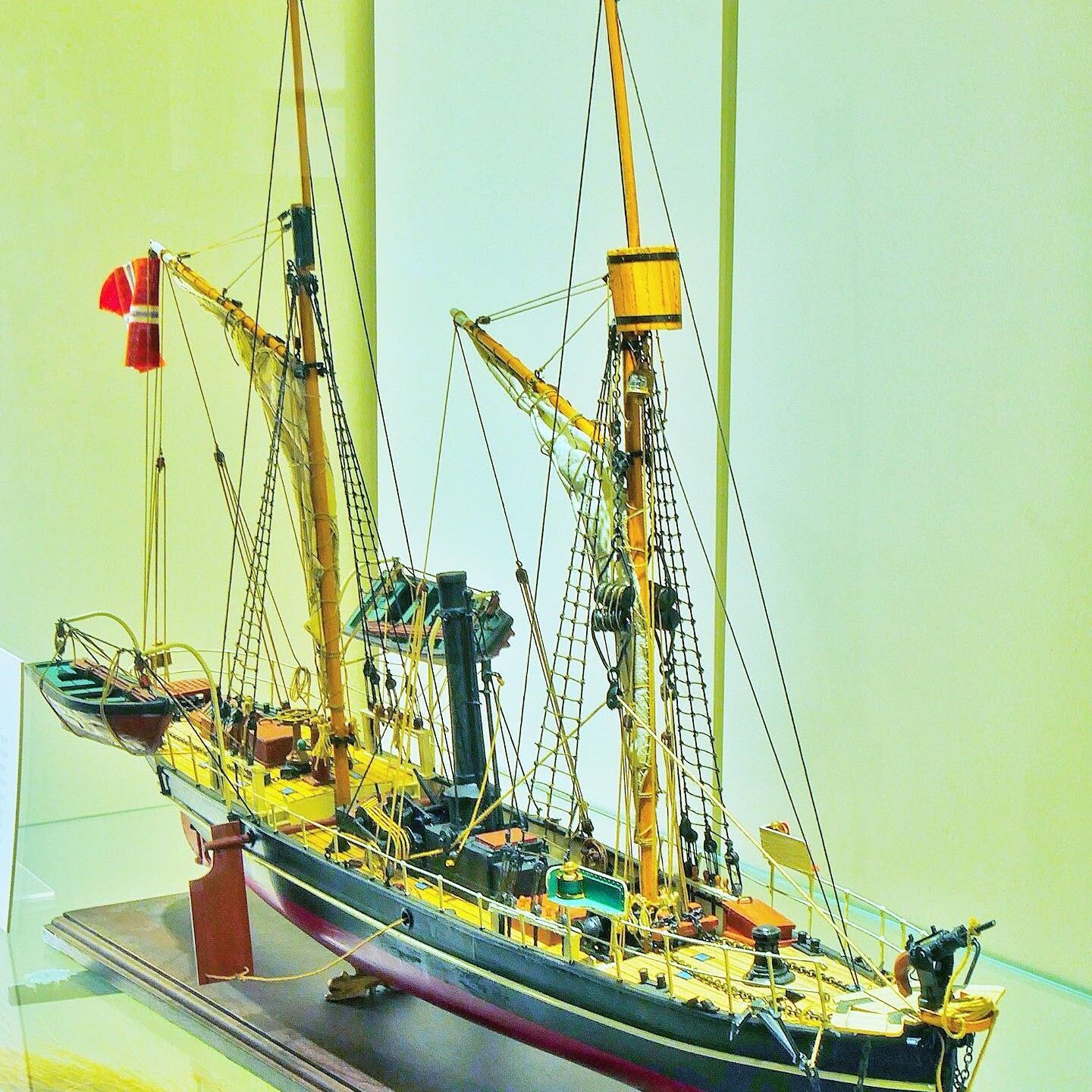 Detailliertes Modell eines historischen Segelschiffs mit zwei Masten, Takelage, Beibooten und gelb-rotem Deck in Vitrine.