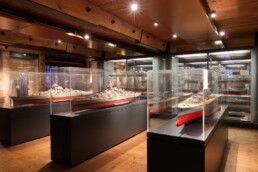 Innenaufnahme eines Museums mit mehreren Glasvitrinen auf dunklen Podesten, in denen detaillierte Schiffsmodelle (Schlachtschiffe) mit rot gestrichenen Unterseiten unter warmer Holzdeckenbeleuchtung ausgestellt sind.