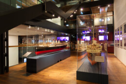 Innenansicht eines Museums: zwei große, detaillierte Schiffsmodelle in gläsernen Vitrinen auf warmem Holzfußboden, umgeben von moderner Beleuchtung, einer Stahltreppe und Bildschirmen sowie gerahmten Fotos an der Wand.