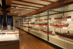 Museumsausstellung in einem Raum mit sichtbaren Deckenbalken und Holzboden; rechts lange Glasvitrinen mit mehreren detaillierten Schiffsmodellen mit roten Rümpfen, links flache Vitrinen mit Dokumenten.