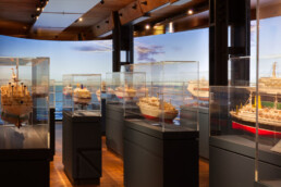 Museumsausstellung mit mehreren detaillierten Schiffsmodellen in Glasvitrinen auf dunklen Sockeln, Holzfußboden und -decke sowie einem großformatigen Panoramahintergrund mit Meer und Himmel.