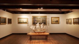 Modell eines historischen Segelschiffs in einer Glasvitrine auf einem Holztisch, zentral in einem Museumssaal mit Holzfußboden und -decke, umgeben von gerahmten Seekunstgemälden an den Wänden.
